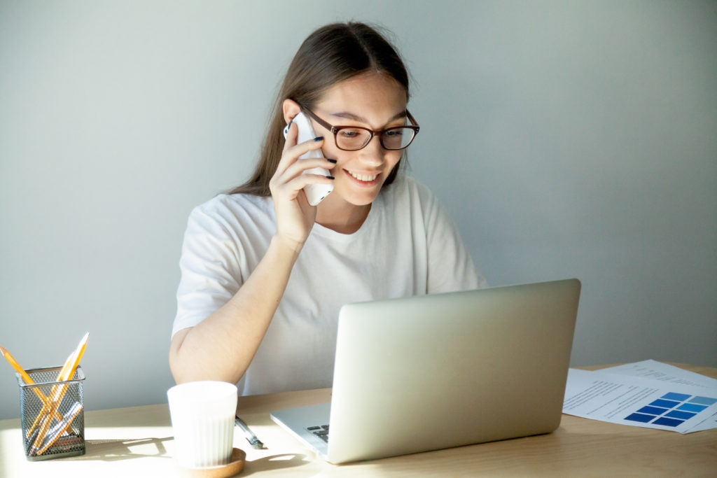 Millennial woman in glasses talking on mobile and using laptop teletrabajo